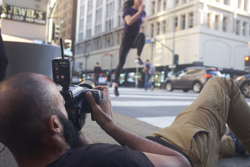 Photographer holding camera while seated on urban street with city buildings and pedestrians in background.