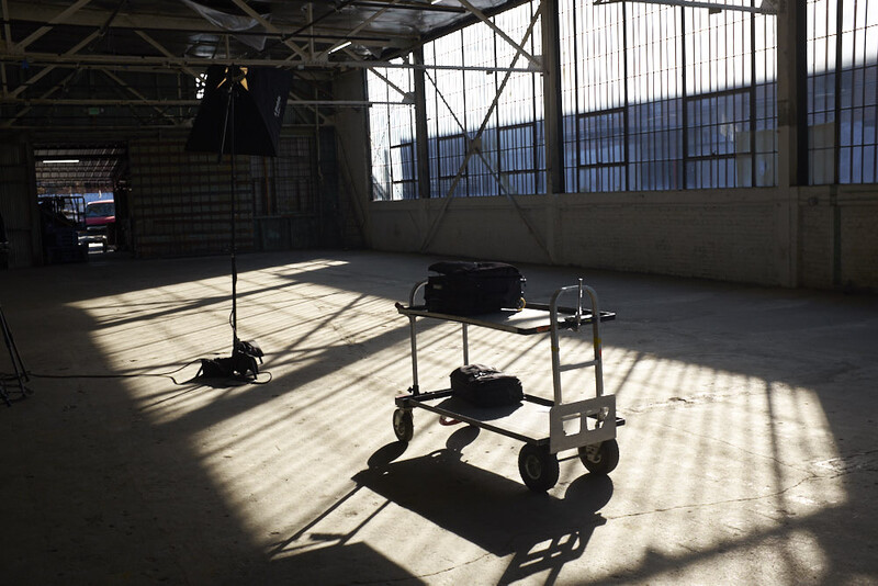 Industrial studio interior with overhead lighting rigs and equipment carts casting sharp shadows on concrete floor.