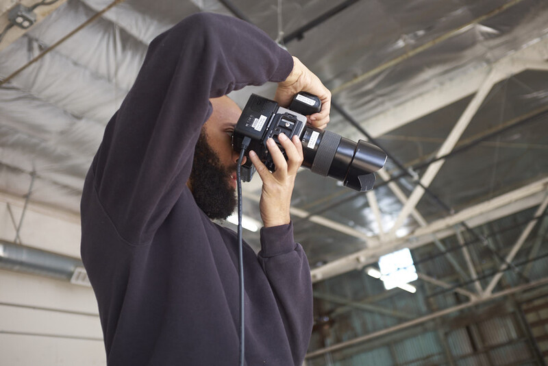 Bearded photographer in dark hoodie holding vintage film camera up to face in industrial warehouse space.