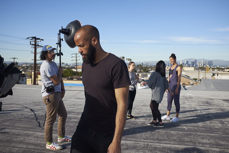 Man holding professional cinema camera on rooftop with city skyline and crew in background.