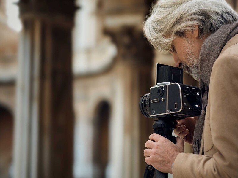 Photographer holding a medium format film camera up to their face, with blurred classical architecture in the background.