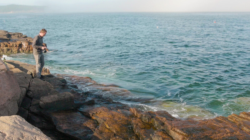 Fisherman standing on rocky coastline casting a line into calm turquoise water.