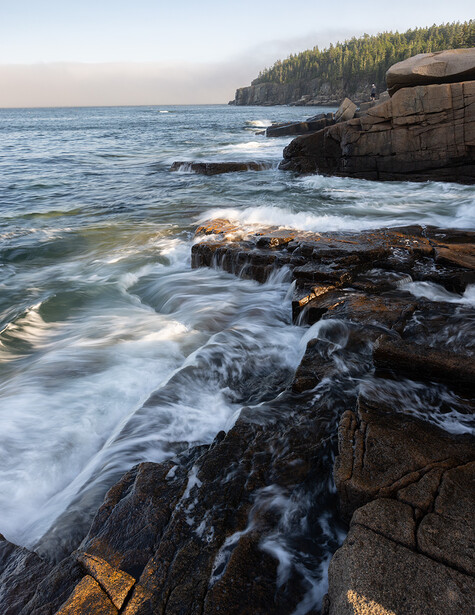 Rocky coastline with crashing waves and forested headland at twilight.