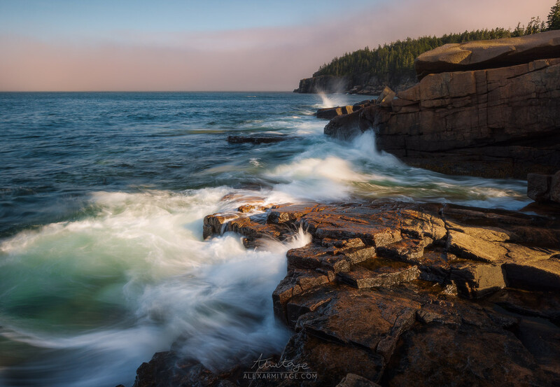 Waves crash against rocky shoreline at dusk with forested cliffs in the background.