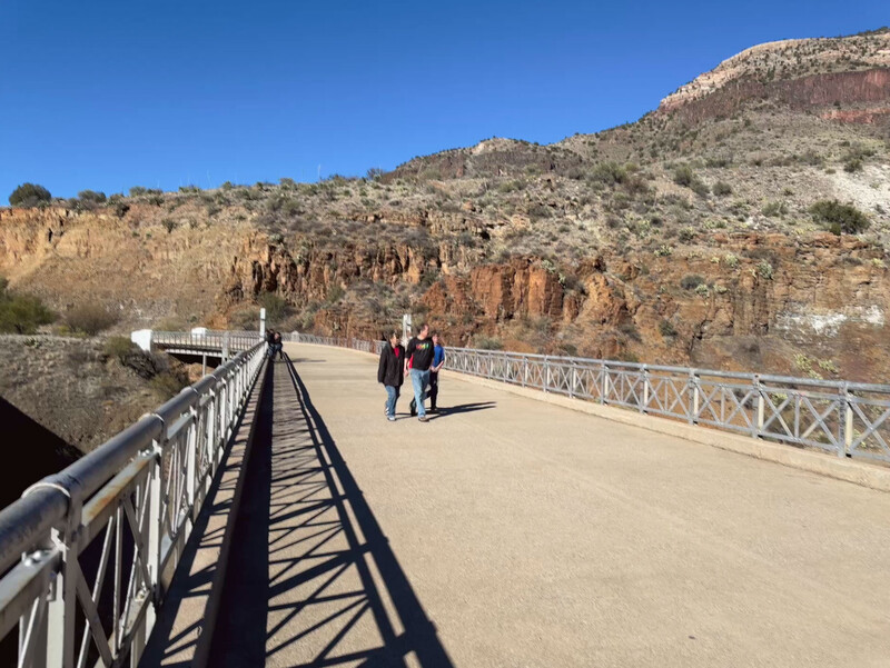 People walking across a metal footbridge spanning a canyon with layered red rock formations and desert vegetation.