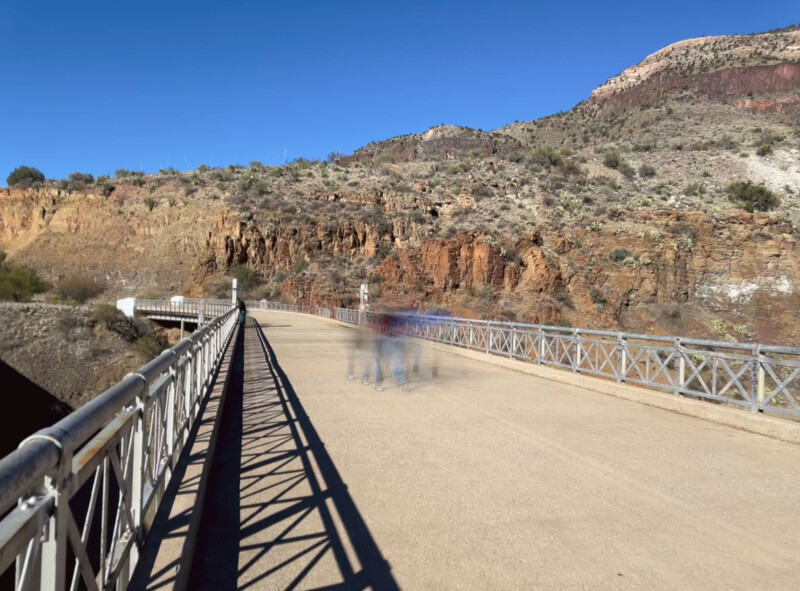 Long pedestrian bridge with metal railings spanning across an arid canyon landscape with layered rock formations.