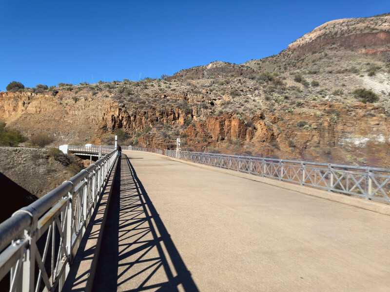 Modern metal bridge spanning across a desert canyon with stratified rock walls beneath clear blue sky.