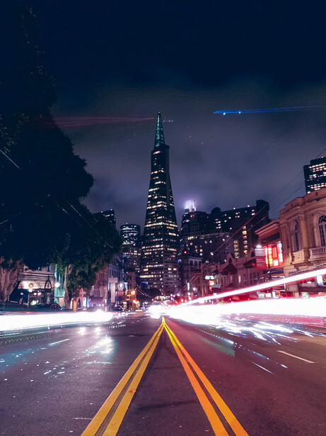 City street at night with yellow road lines and light trails from traffic, tall illuminated skyscraper in background under dramatic cloudy sky.