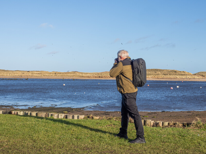 Photographer standing on grass facing a coastal estuary, holding camera to face with backpack on shoulder.