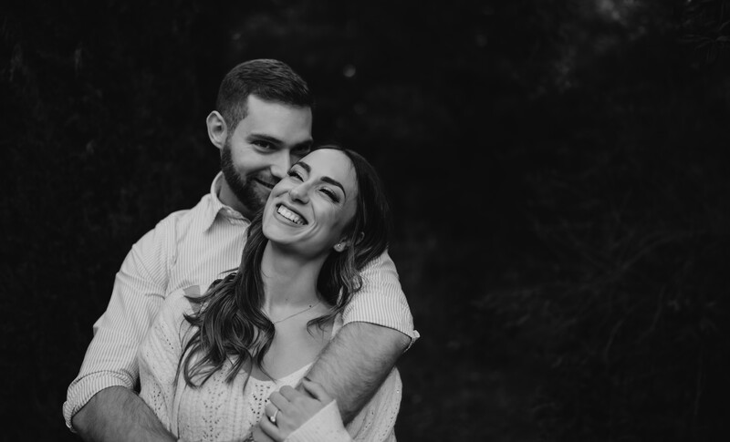 Man embracing woman from behind, both smiling at camera in black and white portrait.