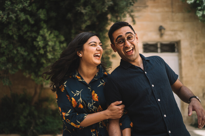 Two people laughing together outdoors against a weathered stone building with ivy.