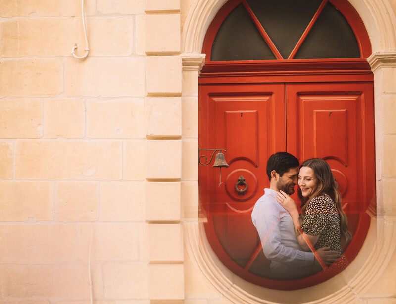Couple embracing in front of a bright red arched doorway set into a cream-colored stone building.