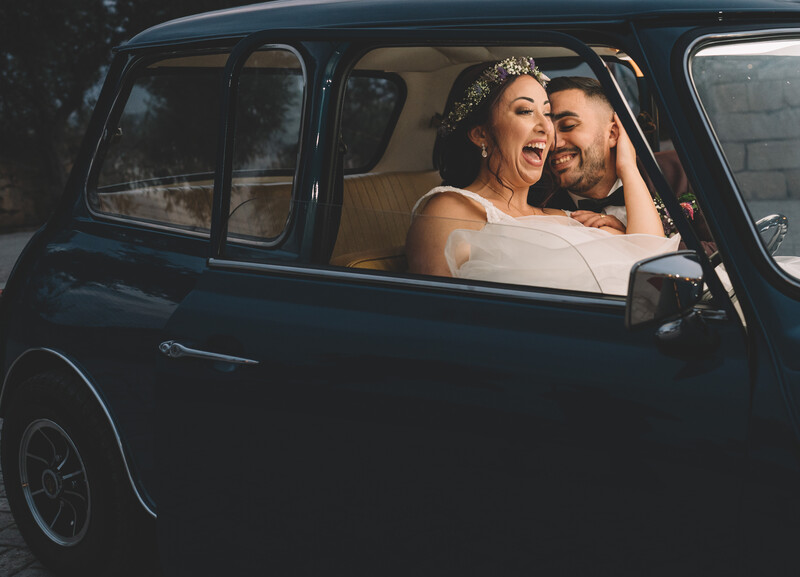Bride and groom smiling through the window of a vintage dark blue car.
