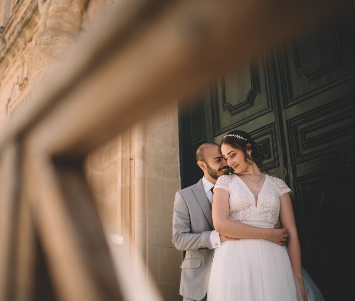 Groom in gray suit kissing bride's cheek as she smiles, both standing against an old stone building with a dark wooden door.