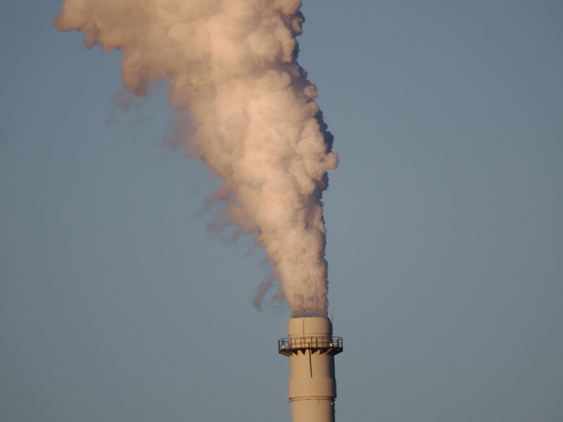 Industrial smokestack emitting thick white and gray smoke against clear blue sky.