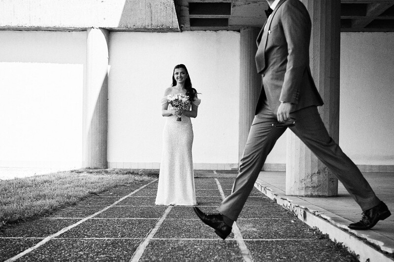Bride in white dress holding bouquet stands still while groom in suit walks past on geometric floor pattern.