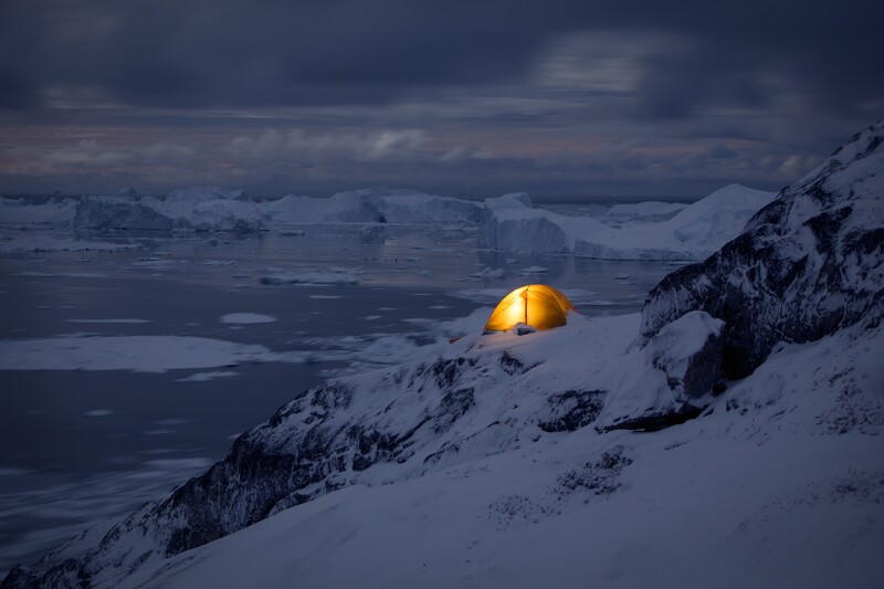 Illuminated yellow tent glowing on a snowy mountainside at twilight overlooking a frozen landscape.