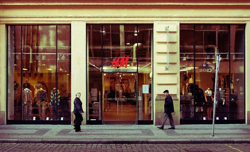 Storefront with large windows displaying mannequins and merchandise, pedestrians walking past on a brick street.