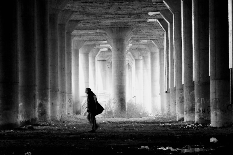 Silhouetted figure standing beneath a concrete overpass with dramatic light streaming through structural columns.