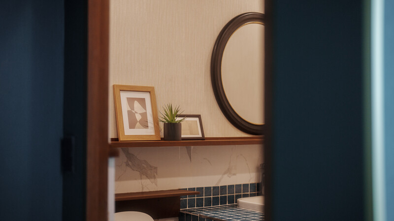 Framed artwork and small potted plant on wooden shelf beside round mirror against warm beige wall.
