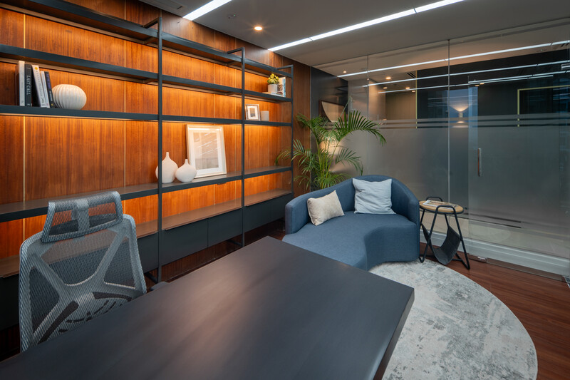Modern office lounge with wooden shelving wall, blue seating, and glass partition dividing the space.