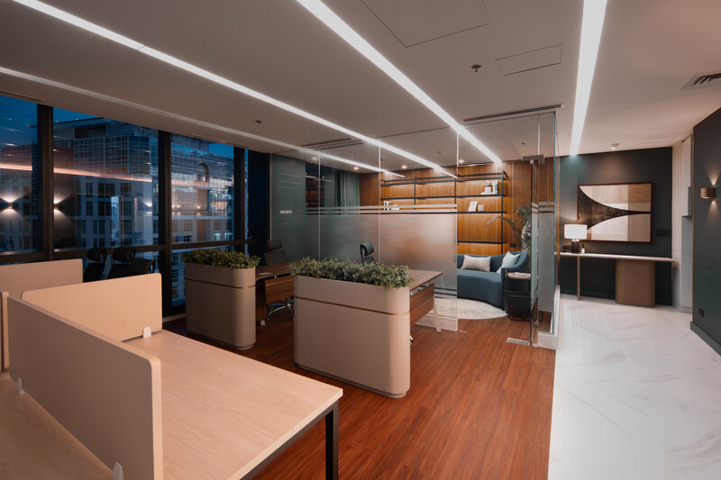 Modern office interior with linear ceiling lights, wooden flooring, and floor-to-ceiling windows overlooking a city skyline at dusk.