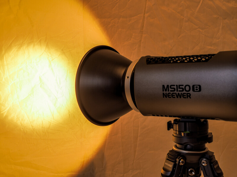 Neewer MS150B monolight mounted on tripod, illuminating a textured backdrop with warm golden light.