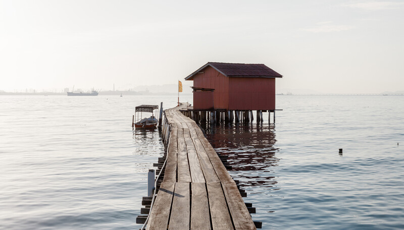 Wooden boardwalk extending toward a red boathouse on stilts in calm water under overcast sky.