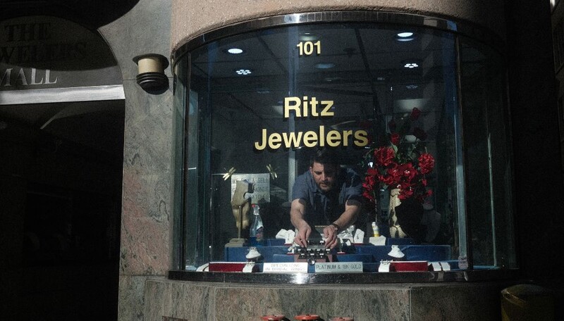 Jeweler working at storefront window display with flowers and merchandise visible behind glass.
