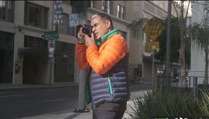Photographer holding a camera to his face on an urban street, wearing a bright orange and gray puffy jacket.