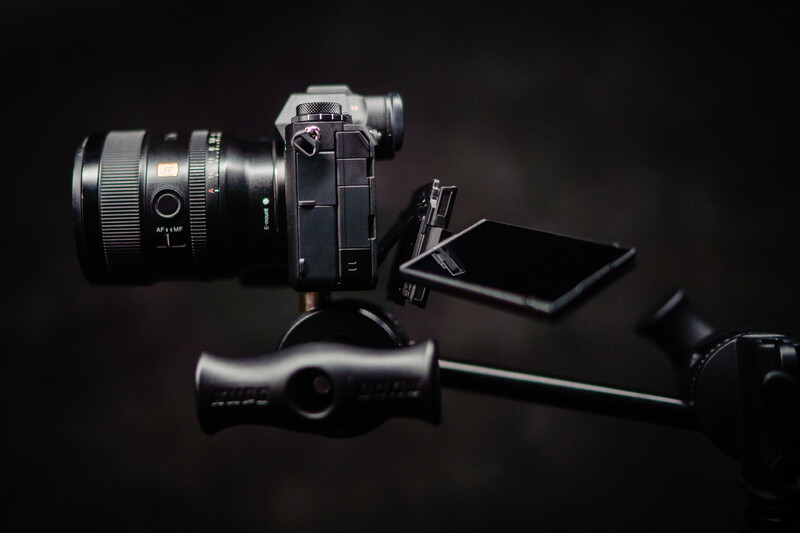 Mirrorless camera with attached lens, tripod head, smartphone, and accessories arranged on dark background.
