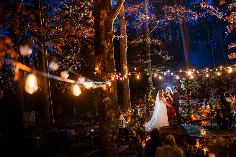 Bride and groom standing together under string lights in a wooded outdoor venue at night.