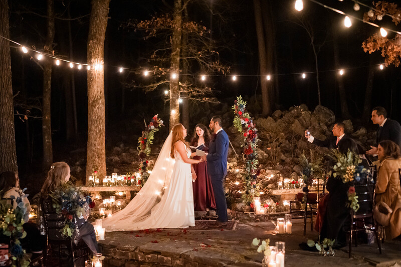 Bride and groom exchange vows at an outdoor wedding ceremony illuminated by string lights and candles.