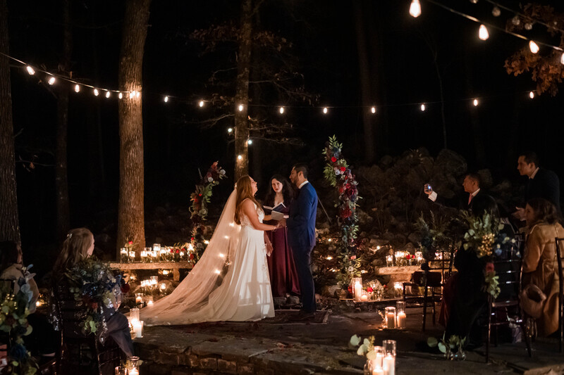 Bride and groom exchange vows at an outdoor nighttime wedding ceremony beneath string lights and trees.