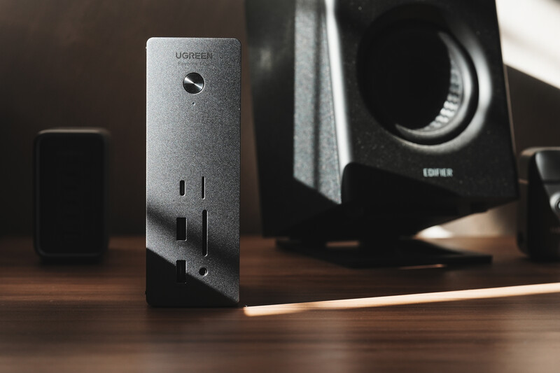 Ugreen power adapter and speaker on wooden desk with warm backlighting.