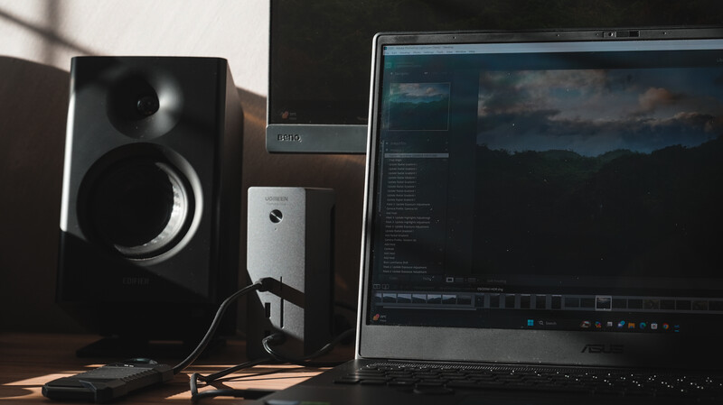 Photo editing workspace with studio monitor, powered speaker, and laptop displaying landscape image in post-processing software.