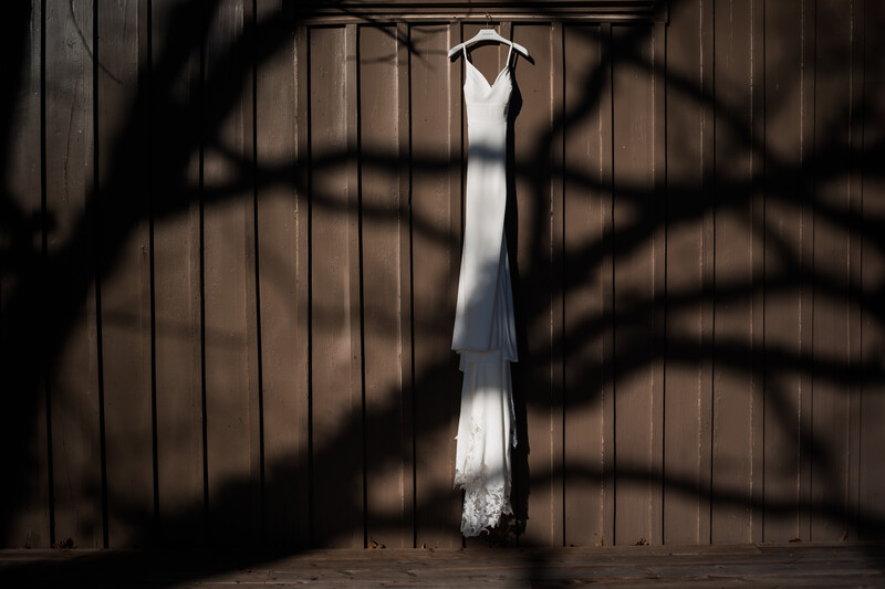 White wedding dress hanging from wooden beam with dramatic tree branch shadows cast across wooden wall.