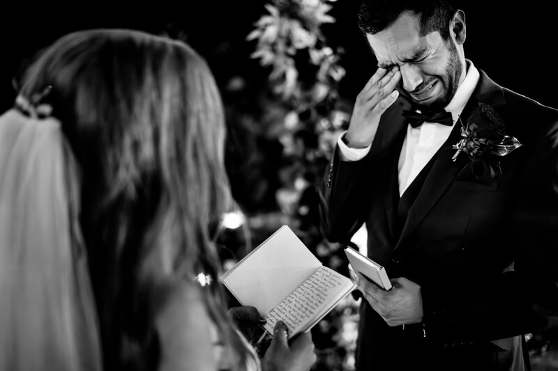 Groom reading vows during wedding ceremony while bride listens in black and white.