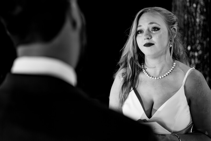 Woman in white wrap dress and pearl necklace gazes at man in tuxedo in black and white portrait.