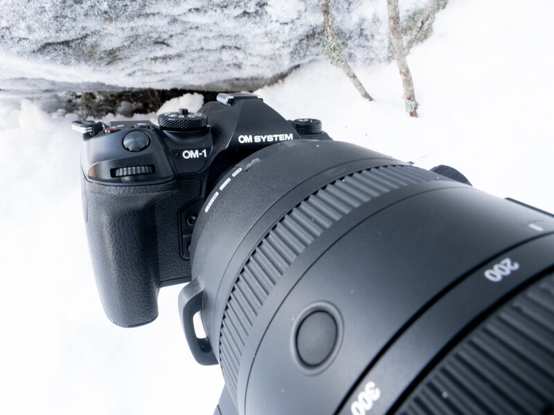 OM system camera with telephoto lens mounted on snowy surface beneath ice formation.
