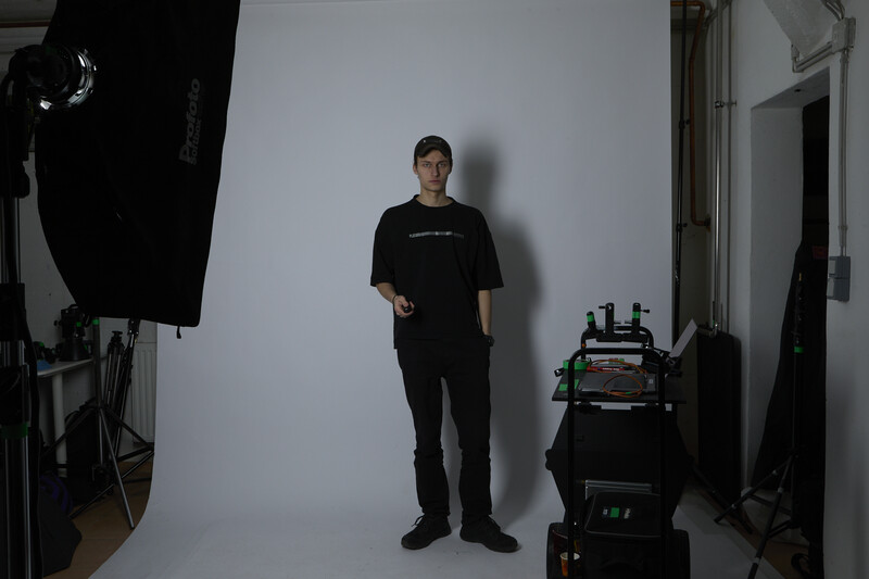 Man in black clothing standing against white backdrop in studio with professional lighting equipment visible.