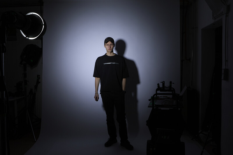 Man in black shirt standing in studio lit by overhead key light with dramatic shadow on backdrop.