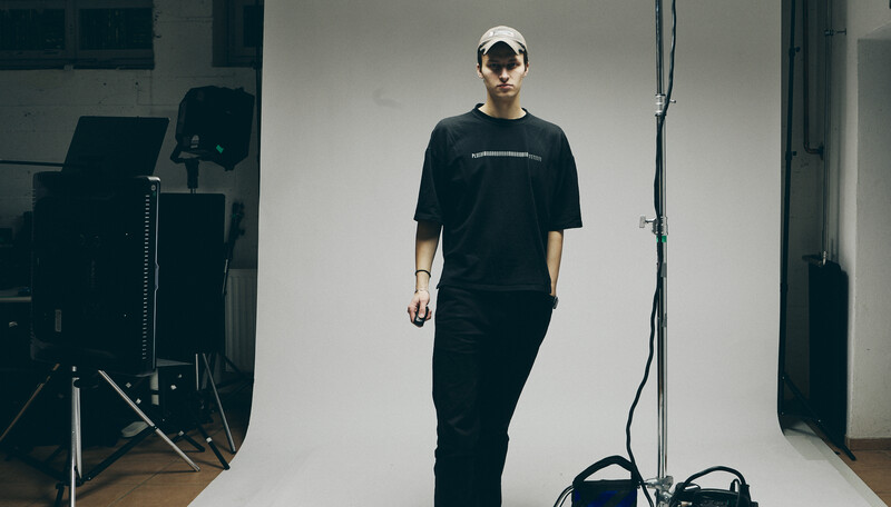 Person in black t-shirt and pants standing in white studio with professional lighting setup and equipment visible.