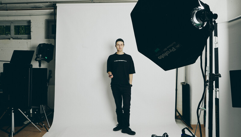 Man in black clothing standing centered against white backdrop in professional studio with large softbox lighting.