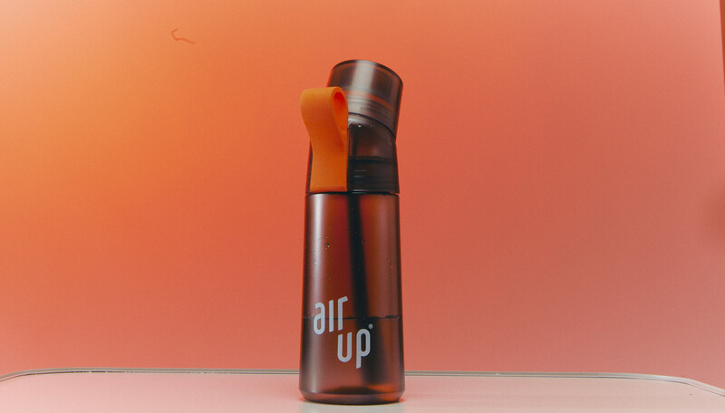 Brown spray bottle with orange atomizer nozzle against warm peachy-orange background.