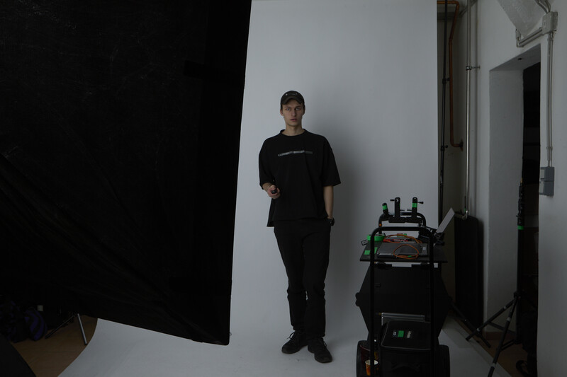 Person in black clothing standing against white backdrop with studio lighting equipment visible on right side.