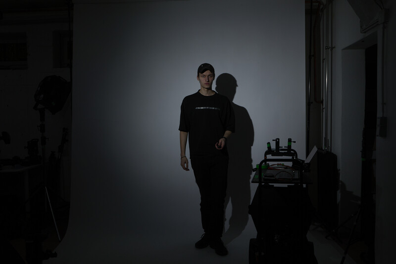 Two silhouetted figures standing in a minimalist white studio space with studio lighting equipment visible.