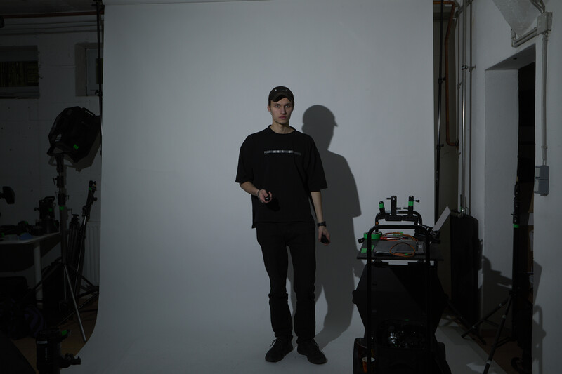 Person in black clothing standing against white backdrop in studio with visible shadow and equipment.
