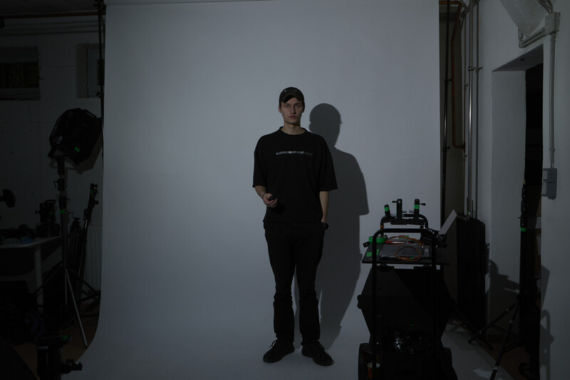Person in black clothing standing against white backdrop in studio with visible lighting equipment.