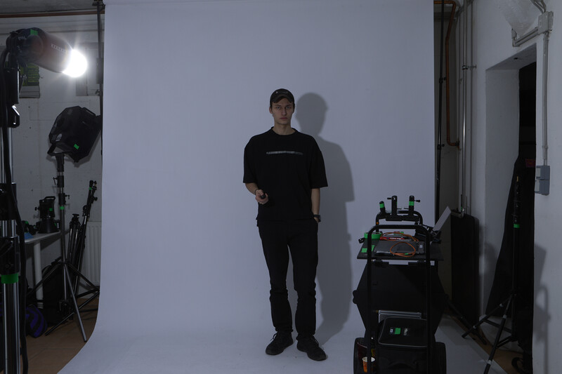 Man in black clothing standing in a photography studio with professional lighting equipment and a white backdrop.